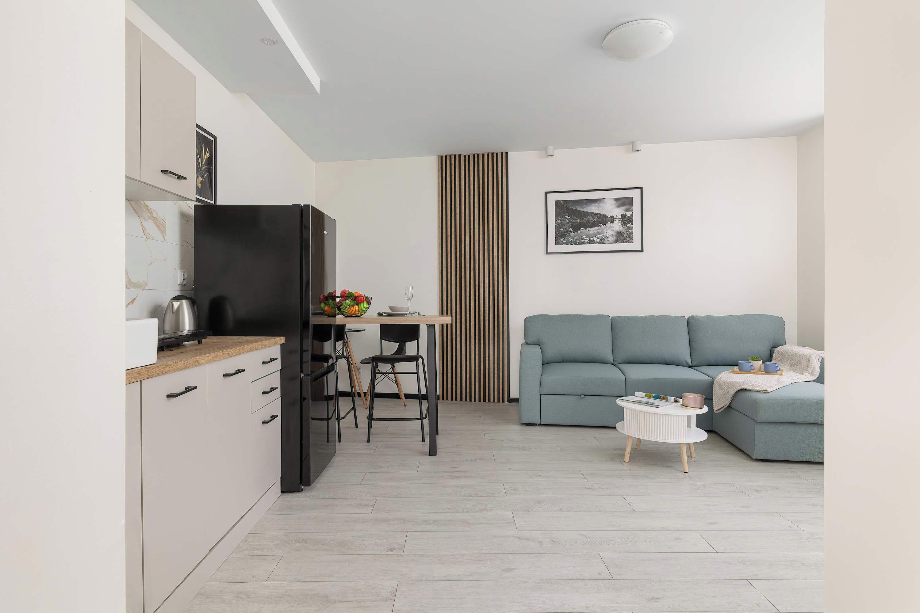 A modern kitchen with a black refrigerator, wooden cabinets, and a marble backsplash. There is a dining area with a table, chairs, and a fruit bowl. A grey sofa and a small white table with a magazine are in the living area.
