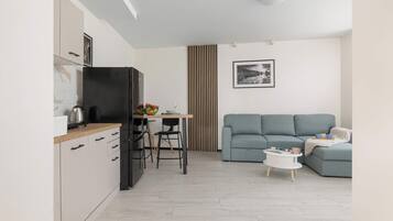 A modern kitchen with a black refrigerator, wooden cabinets, and a marble backsplash. There is a dining area with a table, chairs, and a fruit bowl. A grey sofa and a small white table with a magazine are in the living area.