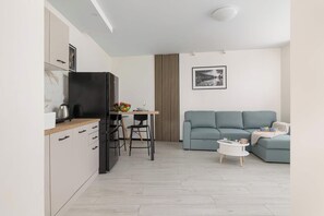 Una cocina moderna con un refrigerador negro, armarios de madera y un protector contra salpicaduras de mármol. Hay una zona de comedor con una mesa, sillas y un cuenco de frutas. Un sofá gris y una pequeña mesa blanca con una revista están en la sala de estar.