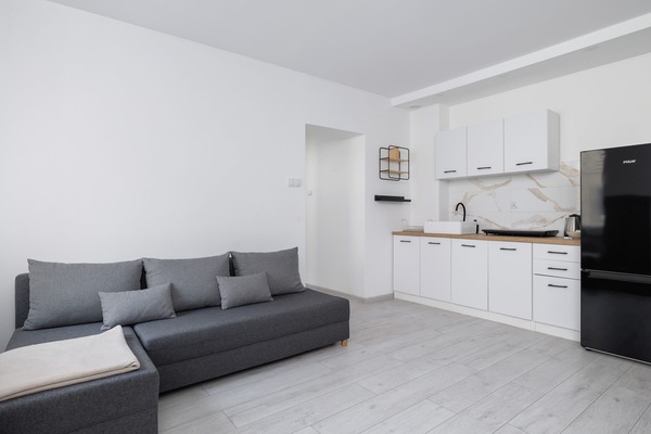 Bright and spacious living area with a grey sofa, open kitchen, and natural light from large windows.