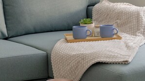 Un salon moderne avec un canapé gris, un plaid blanc à texture intéressante et une petite table sur laquelle se trouvent une bougie et un plateau avec deux tasses bleues.