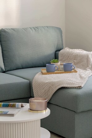 A modern living room with a grey sofa, a white textured throw blanket, and a small table with a candle and a tray holding two blue mugs.