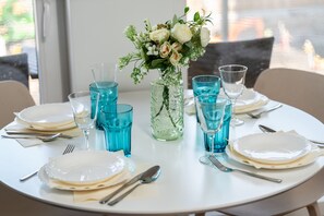Ein runder Esstisch ist mit weißen Tellern, Besteck und blauem Glas ausgestattet, dazu ein Blumenstrauß.