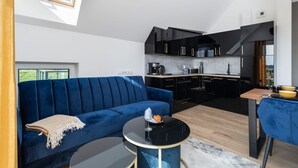 A modern living room with a blue sofa, a small round table, and a kitchen area with black cabinets and a wooden countertop. - Stylish Apartment | Parking, Balcony, Close to the Beach (Sianozety)
