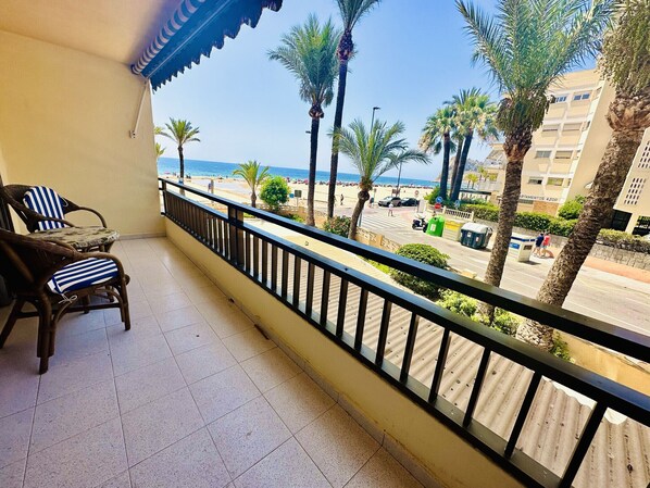 Un balcón con sillas de mimbre, vista a la playa, palmeras y un edificio con la leyenda 'Apartamentos Azor'.