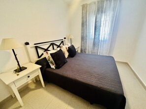 Un dormitorio con una cama, dos mesitas de noche, una lámpara en cada una y una ventana con cortinas.