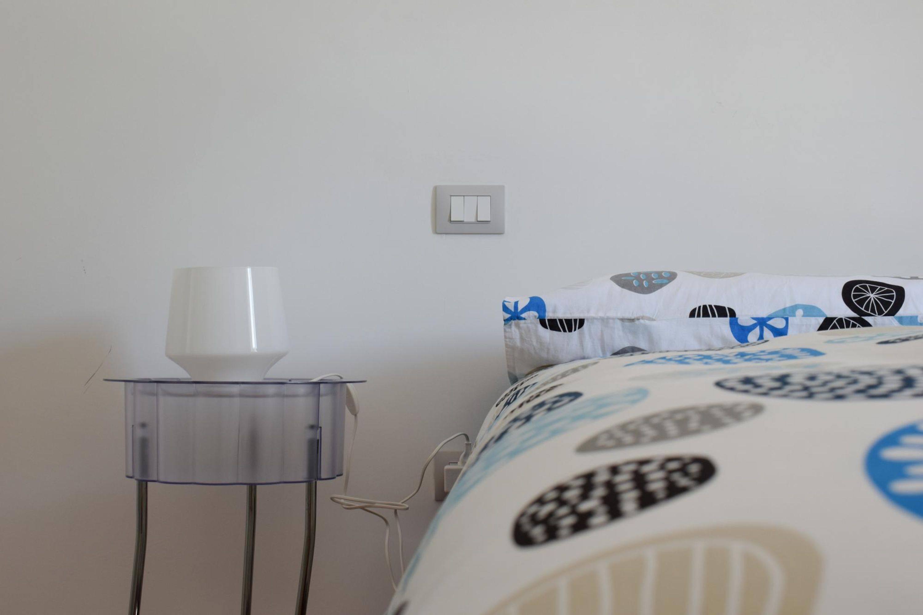 Un lit avec une literie à motifs, une table de chevet avec une lampe blanche et un interrupteur fixé au mur.