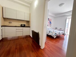 Una habitación compacta con kitchenette, una cama con colcha estampada y una puerta que da a otra habitación.