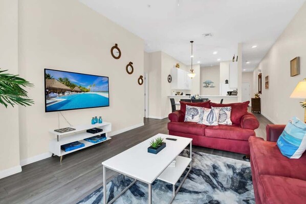 Una sala de estar moderna con una televisión de pantalla plana, un sofá rojo, una mesa de centro blanca y una cocina al fondo.