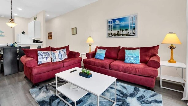 Una sala de estar con dos sofás rojos, una mesa de centro blanca, un tapete azul y un cuadro de una playa.