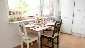 Un comedor con una mesa de madera preparada para cuatro personas, sillas blancas y negras, una ventana con vista a follaje y un refrigerador blanco al fondo.