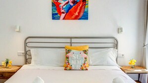 Uma cama bem arrumada com roupa de cama branca e um cobertor amarelo, um travesseiro decorativo com tema tropical e um quadro de sereia acima da cama.