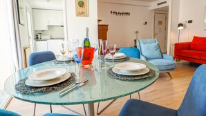 Uma área de jantar com mesa de vidro, cadeiras e um lustre. Cozinha visível ao fundo.