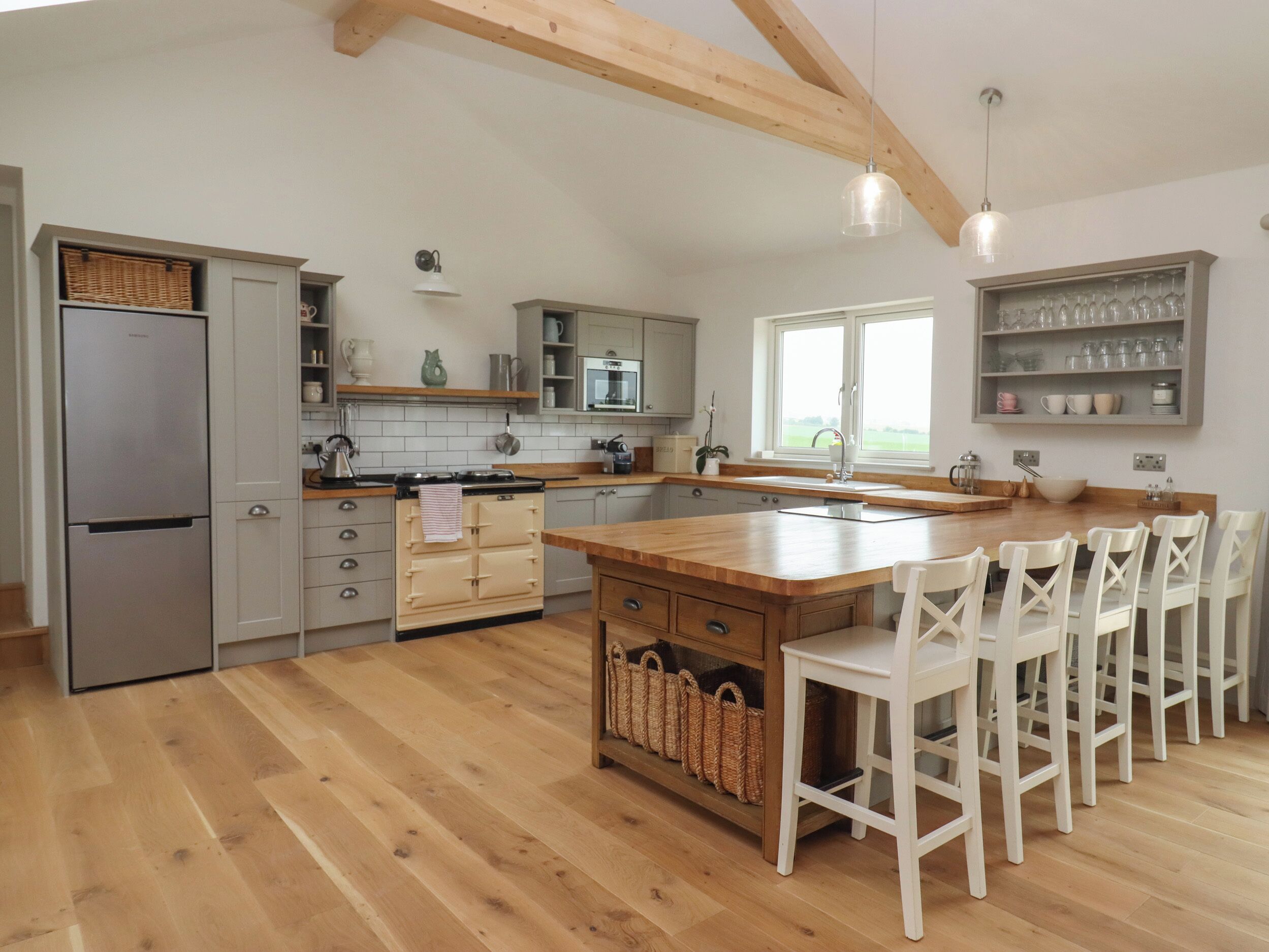 A modern kitchen with wooden flooring, a built-in oven, a large island with bar stools, and a window overlooking the garden.