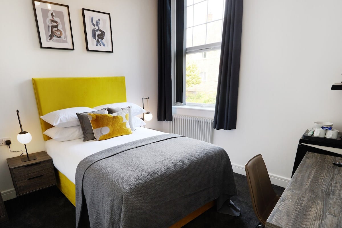 A hotel room with a bed, bedside table, lamp, artwork, window, and a desk with a computer and coffee maker.