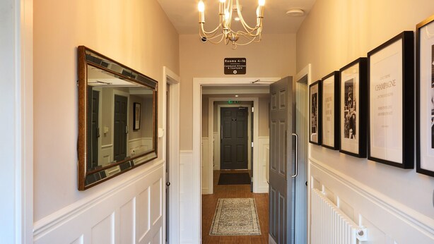 A hallway with white wainscoting, a chandelier, framed pictures, and a sign indicating rooms 4-16. - The Woden Boutique Hotel Walsall M6 J9 (Wednesbury)