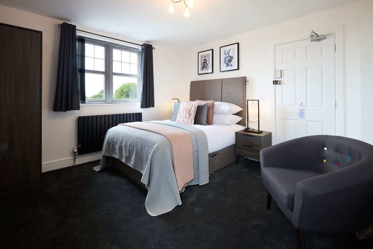A bedroom with a bed, bedside table, chair, window, and artwork on the wall.