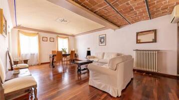 Un ampio soggiorno con soffitto alto, pavimento in legno, un divano bianco, un tavolo da pranzo con sedie e un camino.