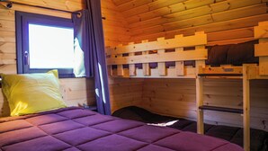 A wooden cabin room with a bed, bunk beds, and a window with curtains.