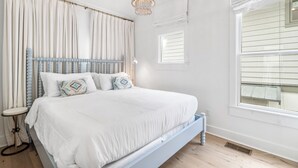 A bedroom with a large bed, a chandelier, a window with blinds, and a bedside table. - Crosswinds Carriage House (Panama City Beach)