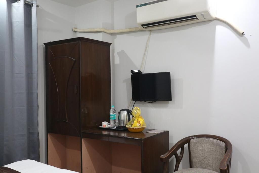 Una habitación con un armario de madera, un escritorio con hervidor, una botella y una cesta. Un televisor está montado en la pared.