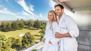 Ein Paar in weißen Gewändern steht auf einem Balkon mit Blick auf eine grüne Landschaft mit Bäumen und einem Gebäude in der Ferne.