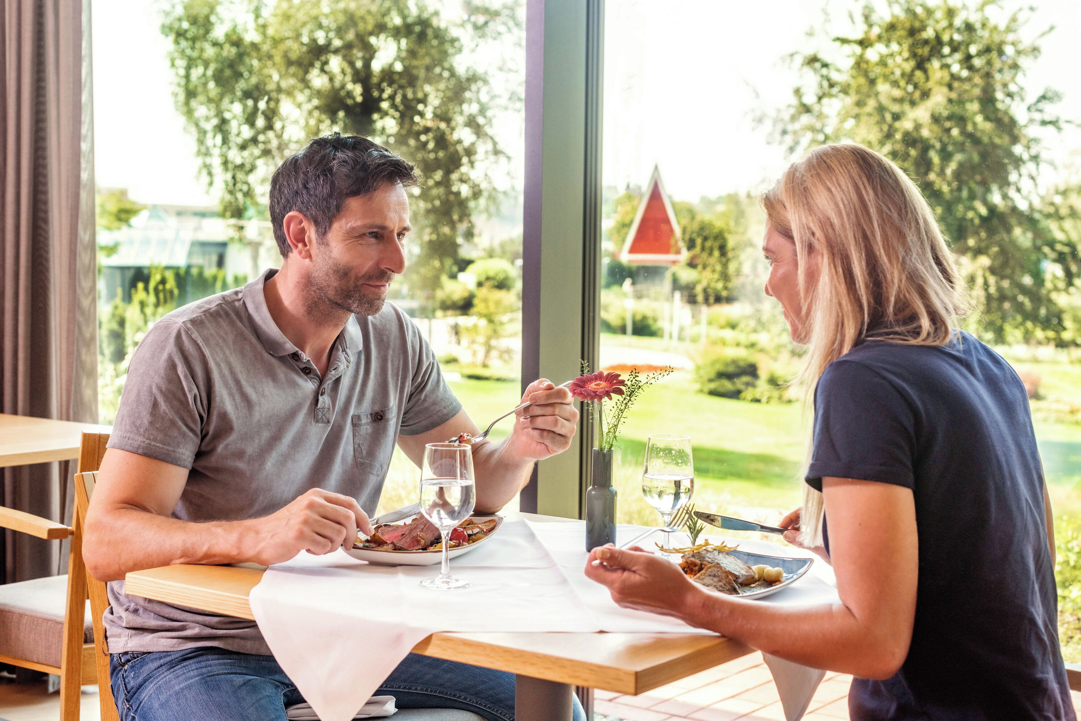 Une table à manger avec deux personnes assises, des assiettes remplies de nourriture et un vase avec des fleurs.