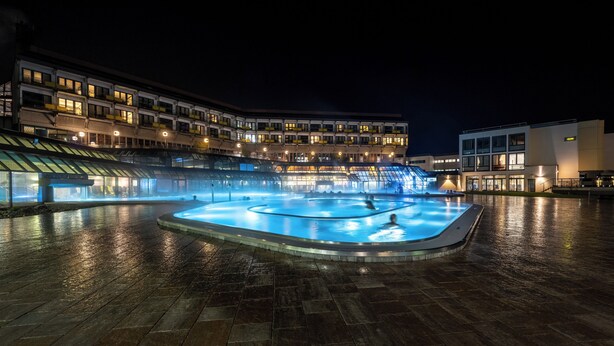 Ein Schwimmbecken mit einer Fontäne, umgeben von einer gefliesten Terrasse, neben einem mehrstöckigen Gebäude mit Balkonen und Fenstern.