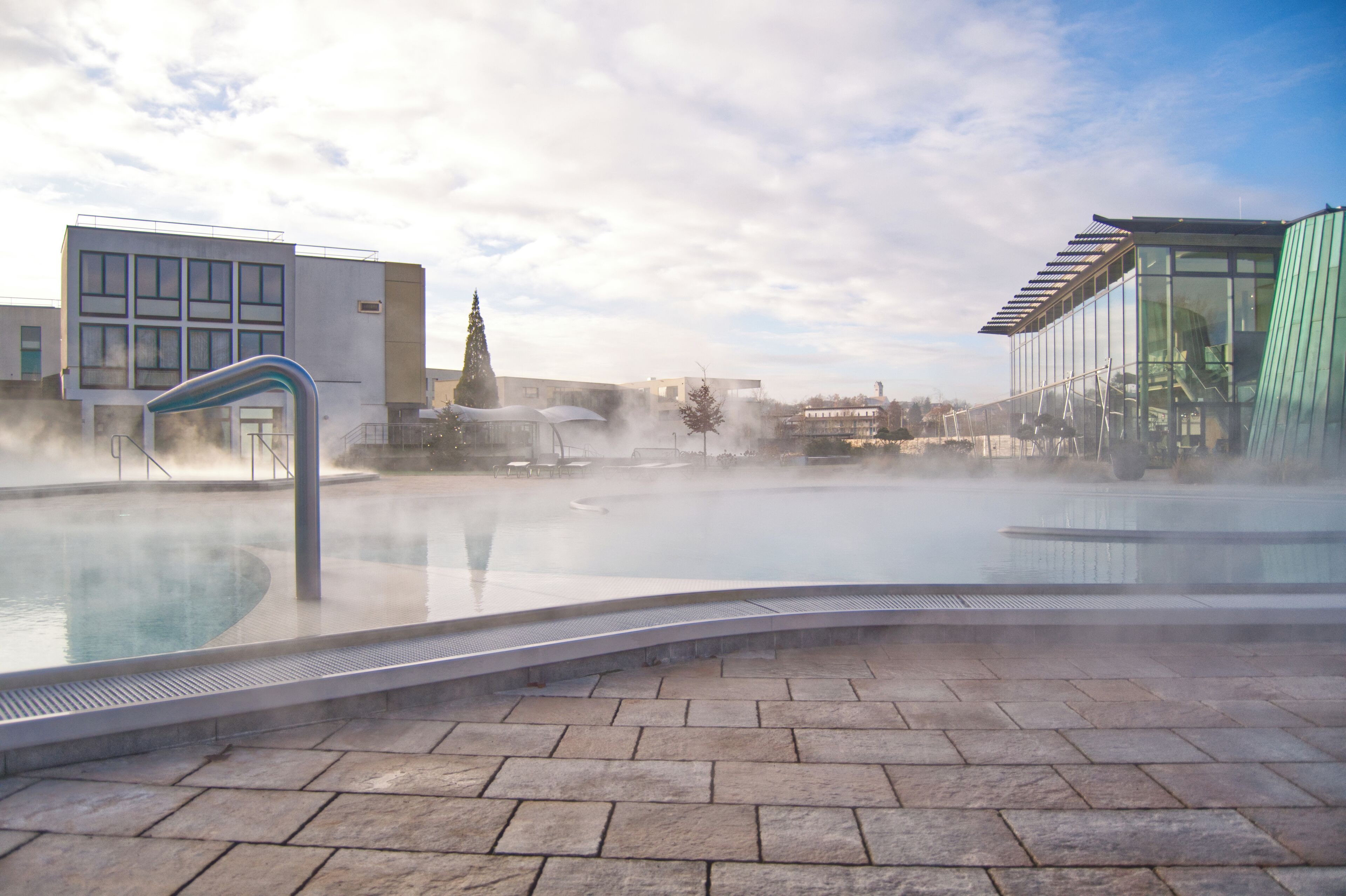 A hot spring pool with steam rising, surrounded by stone tiles and modern buildings.