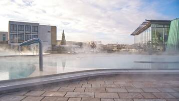 A hot spring pool with steam rising, surrounded by stone tiles and modern buildings.