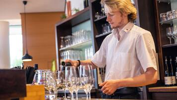 Eine Person gießt ein Getränk in ein Weinglas an einer Bar. Im Hintergrund sind Regale mit Weinflaschen und Gläsern zu sehen.