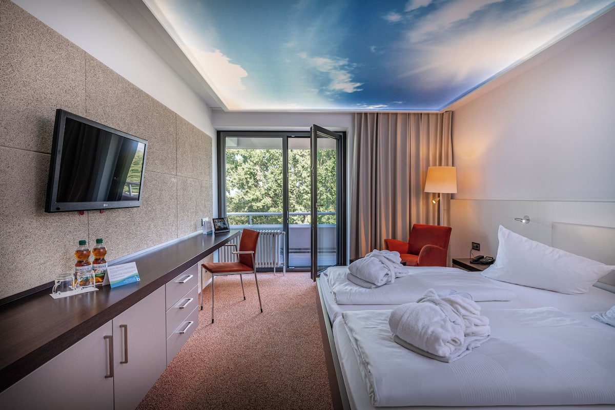 A hotel room with a large bed, a desk with a flat-screen TV, a chair, and a view of the outdoors through a large window.