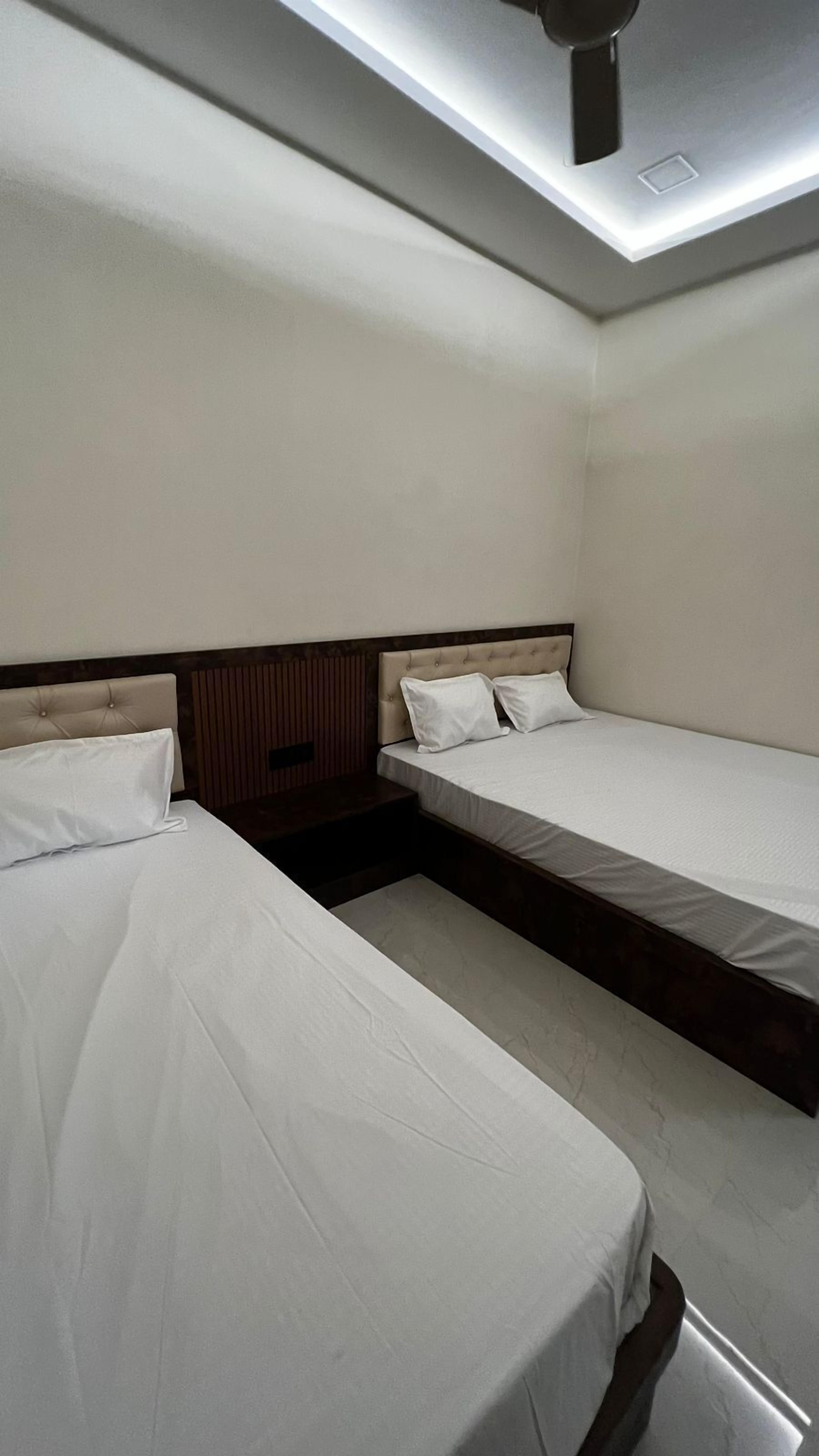 Quarto de hotel com duas camas de solteiro, cabeceira de madeira, mesa de cabeceira pequena e luminária no teto.