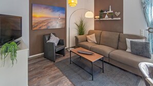 Una sala de estar moderna con un sofá, un sillón y una mesa de centro. Hay un cuadro en la pared, una lámpara de pie y un televisor sobre un mueble.
