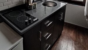 Cocina compacta con encimera negra, salpicadero blanco de azulejos y ventana con vistas a edificios.