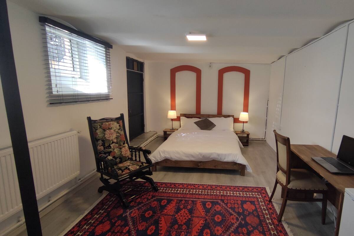 A bedroom with a bed, two bedside tables with lamps, a desk with a laptop, a chair, and a radiator.