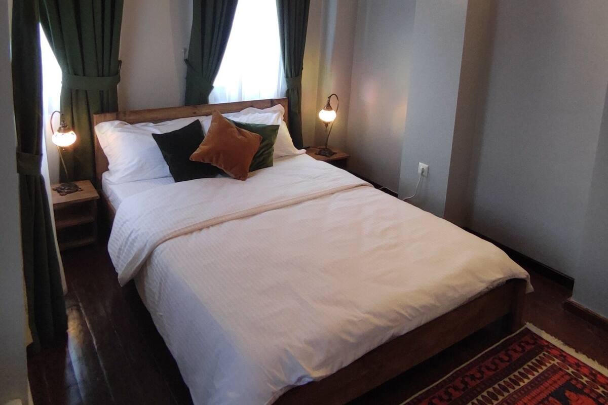 A bedroom with a wooden ceiling, a bed with white bedding, green curtains, and a patterned rug.