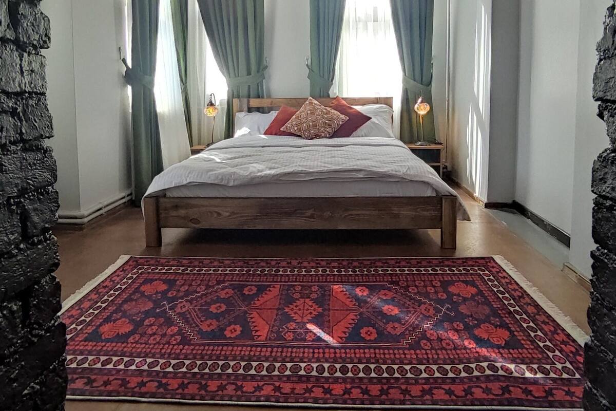 A bedroom with a wooden ceiling, a large bed, green curtains, and a patterned rug.