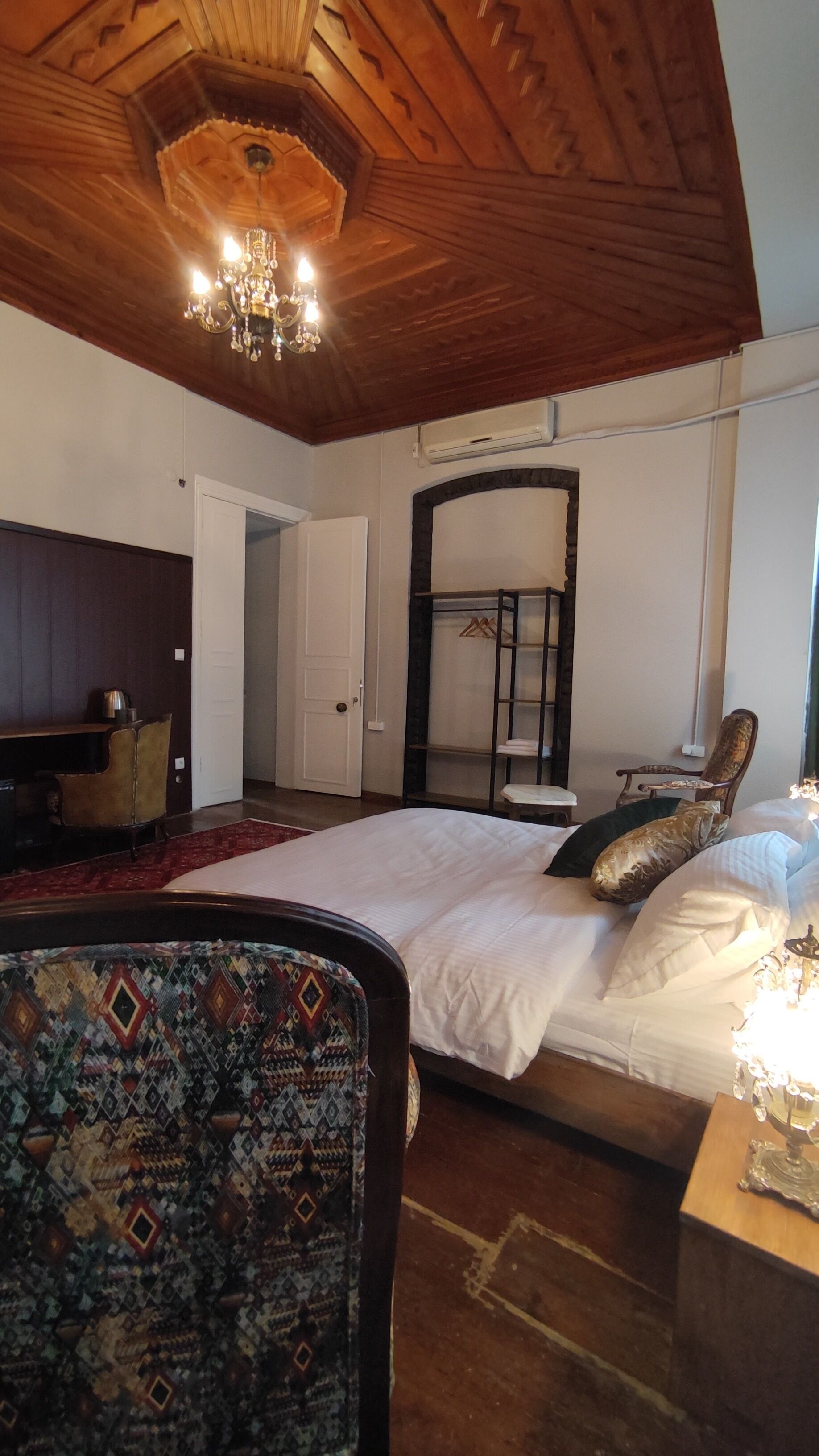 A bedroom with a large bed, a wooden ceiling, a chandelier, a wardrobe, and a chair.