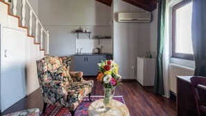 Una sala de estar con un sillón floral, una mesita con un jarrón de flores, una escalera, un área de cocina y un ventanal con cortinas.