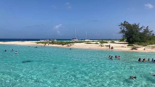 Een strand met helder turquoise water, mensen die zwemmen, en een zeilboot in de verte.