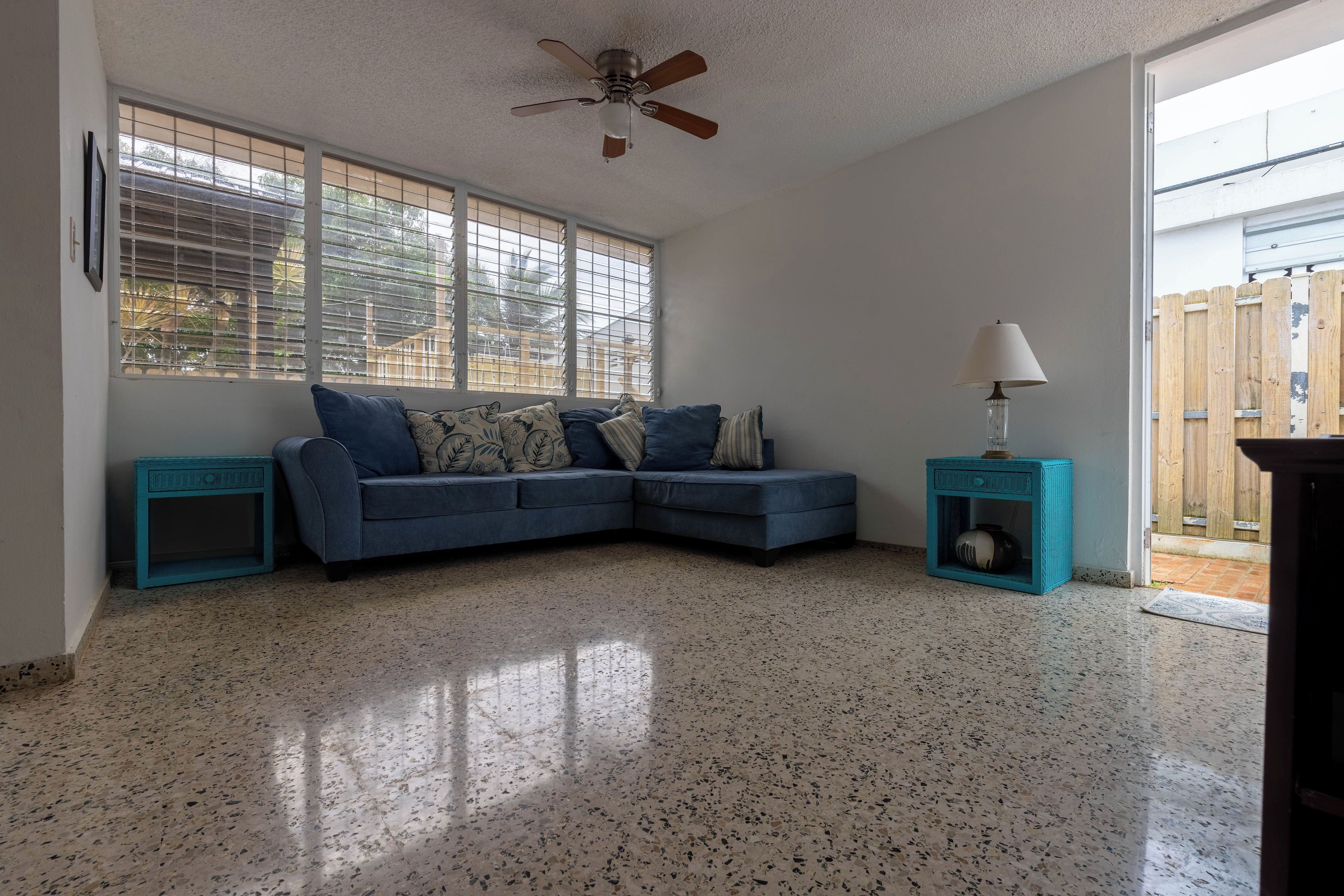 A living room with a blue sectional sofa, a ceiling fan, a small table with a lamp, and a window with blinds.