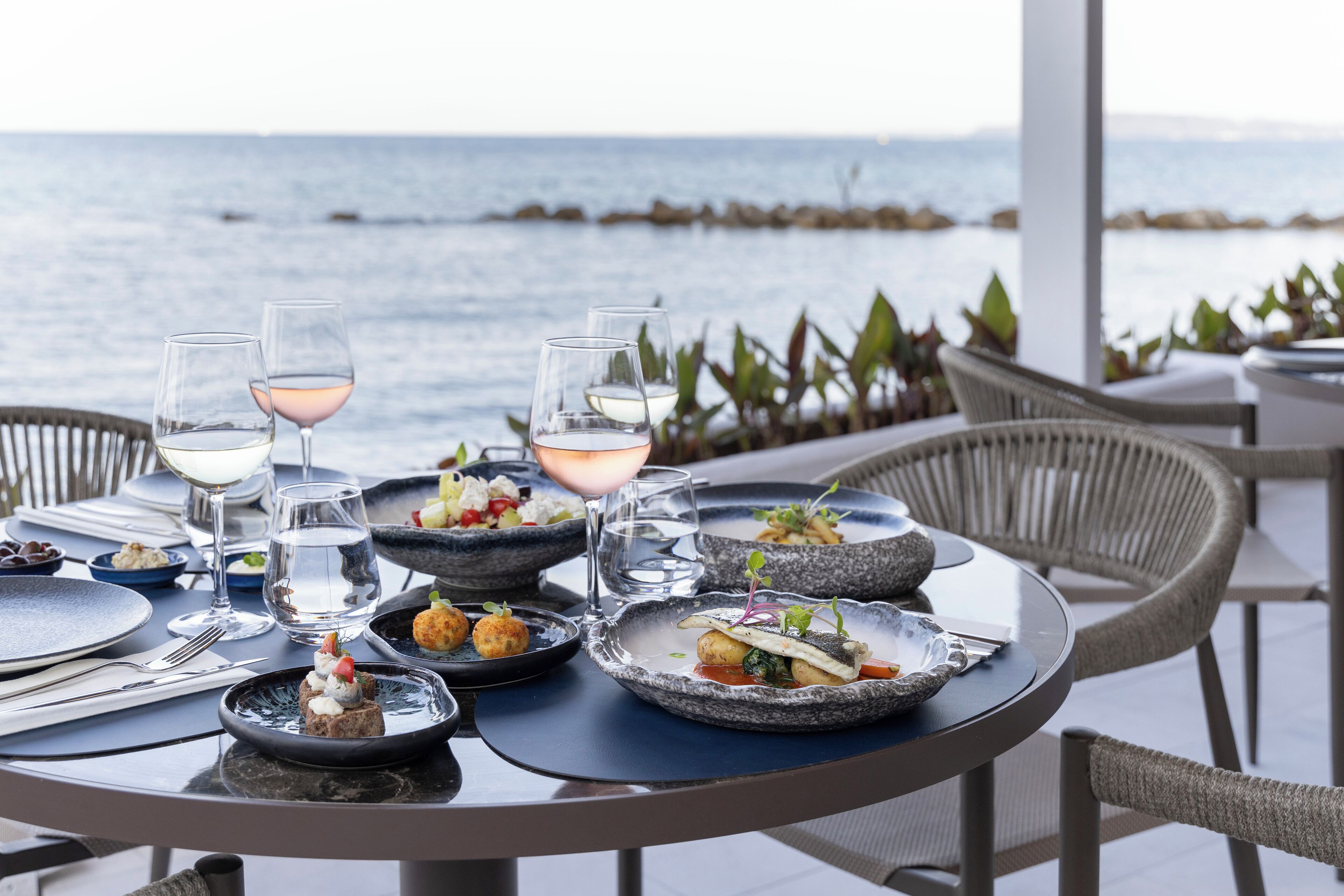 A table set for dining with plates of food, glasses of water and wine, and a view of the sea in the background.