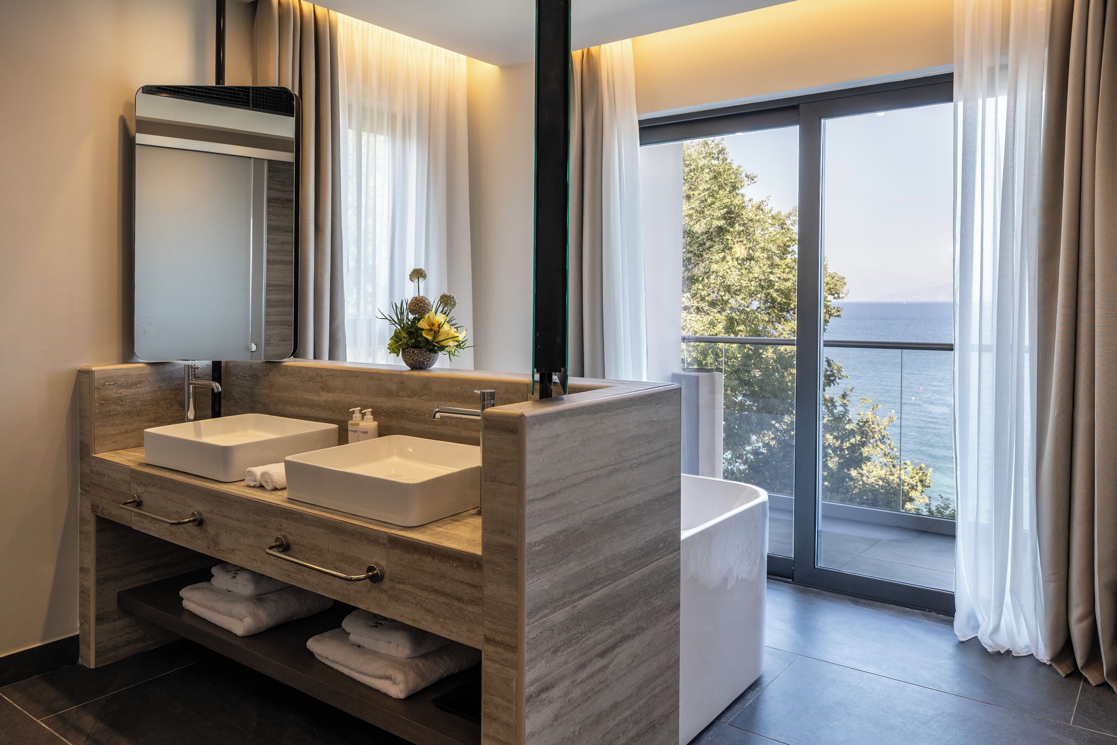 A modern bathroom with two sinks, a large mirror, and a view of the sea through a sliding glass door.
