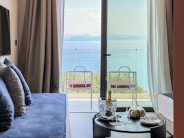 Una habitación con sofá, una mesa con una botella, vasos y fruta, y un balcón con sillas y vista al mar.