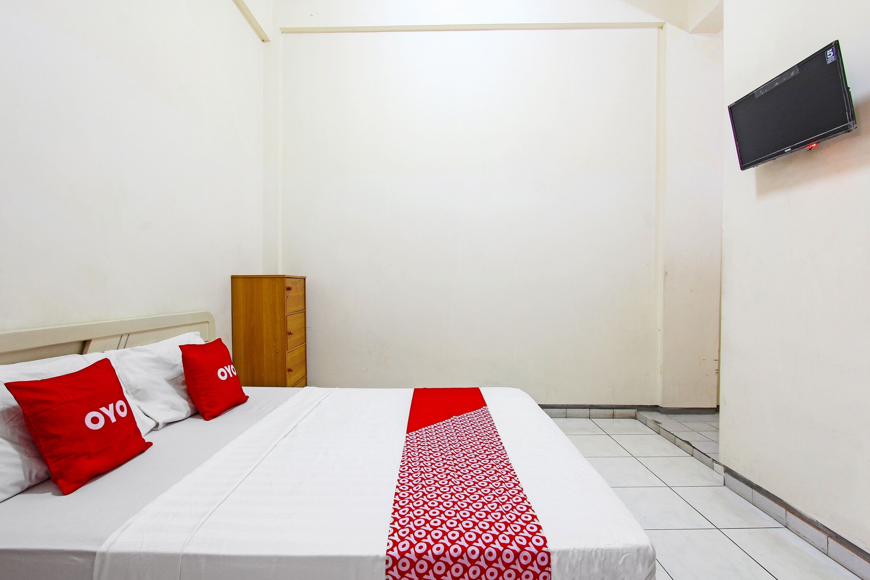 A hotel room with a bed, two red and white pillows, a red and white bedspread, a wooden nightstand, and a flat-screen TV mounted on the wall.