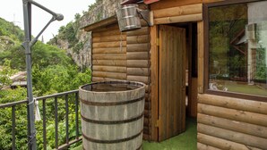 Uma cabana de madeira com um barril grande na varanda, rodeada por vegetação e um cenário montanhoso.