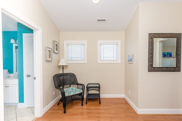 A hallway with a door leading to a bathroom, two windows with blinds, a chair, a small table, and a mirror with an ornate frame. - Sol Mate - 5BR 2nd Row Home with Ocean Views, Pool, Hot Tub & Pet Friendly! (North Topsail Beach)