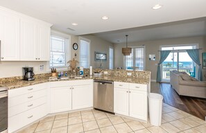 A kitchen with white cabinets, a granite countertop, and a dishwasher. There is a dining area with a view of the ocean. - Sol Mate - 5BR 2nd Row Home with Ocean Views, Pool, Hot Tub & Pet Friendly! (North Topsail Beach)