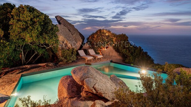 Una piscina con sillas de descanso, rodeada de rocas grandes y árboles, con vistas al océano.